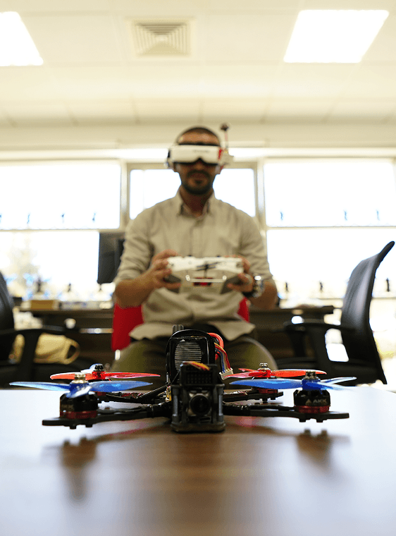 A student wearing FPV goggles operates a drone controller, with a drone in focus on the table in the foreground