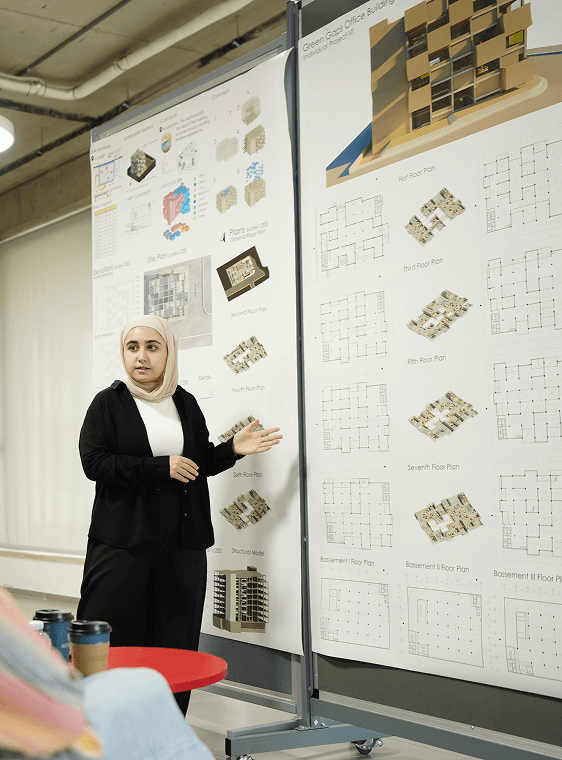 A student presents architectural designs on a board during a discussion, pointing to the visuals